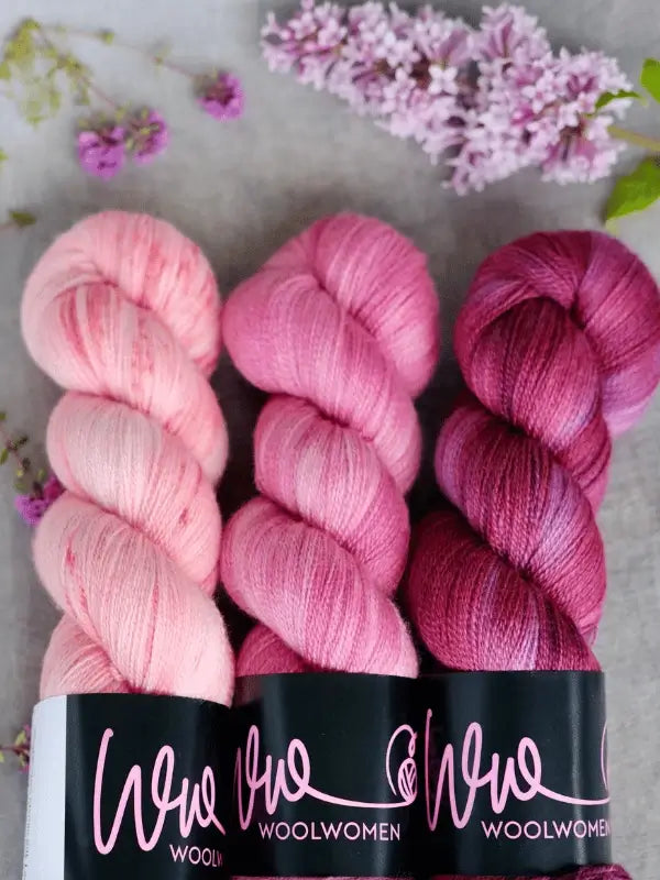 Three skeins of WoolWomen Merino Silk Lace yarn in pink shades are arranged side by side on a grey surface, with purple lilac flowers and petals scattered around them.