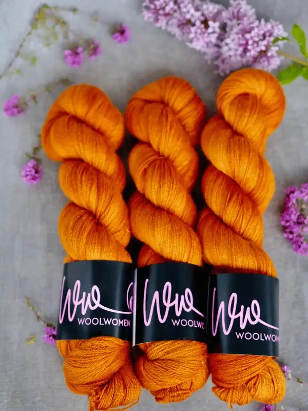 Three skeins of vibrant orange WoolWomen Merino Silk Lace are arranged side by side on a light surface, surrounded by small purple flower sprigs.