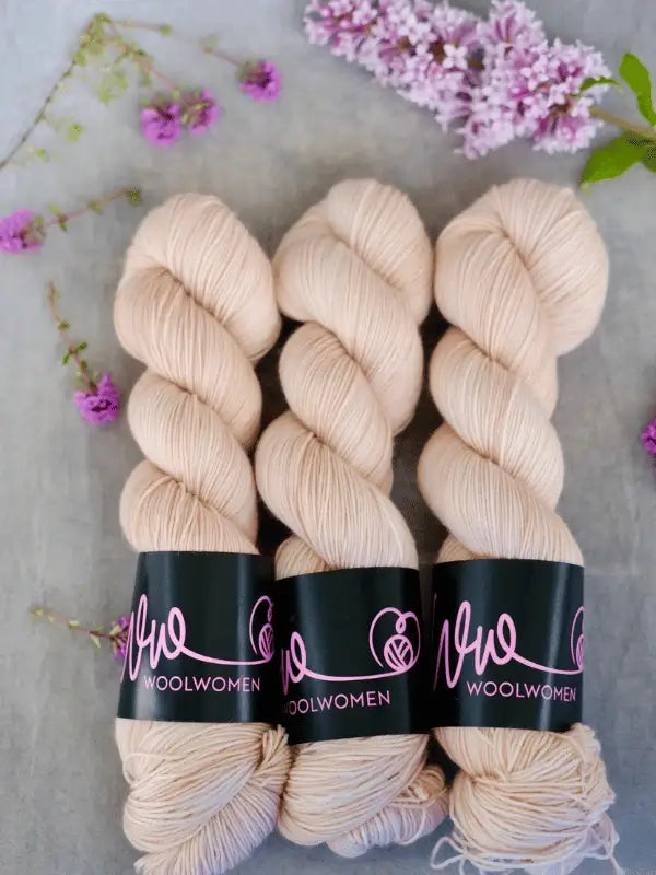 Three skeins of pale beige WoolWomen Merino Silk Lace yarn are arranged side by side, surrounded by small purple flowers and lilac sprigs on a gray background.