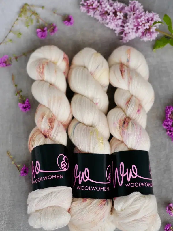Three skeins of WoolWomen Merino Silk Lace yarn with subtle speckles are arranged side by side, surrounded by purple lilac flowers and small blossoms on a light gray background.