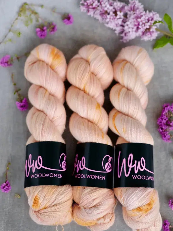 Three skeins of WoolWomen Merino Silk Lace in pale pink with orange speckles are arranged side by side on a gray surface, surrounded by scattered lilac and purple flowers.