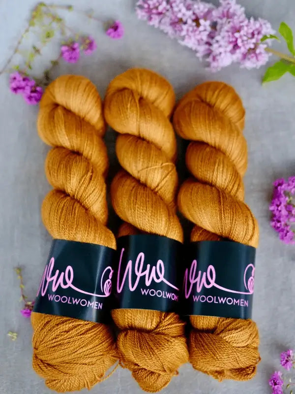 Three skeins of golden-brown WoolWomen Merino Silk Lace are arranged side by side on a light surface, with small clusters of purple flowers scattered around them.