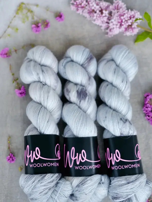 Three skeins of WoolWomen Merino Silk Lace, hand-dyed with light purple speckles and labeled in pink, are displayed side by side on a light background with lilac flower sprigs scattered around them.