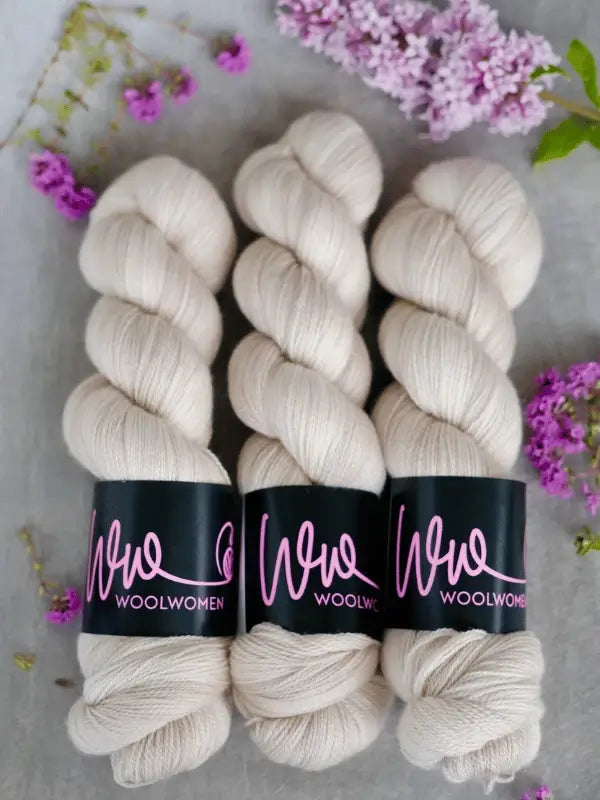 Three skeins of off-white WoolWomen Merino Silk Lace yarn are arranged side by side on a light surface, surrounded by small purple flowers and green leaves.