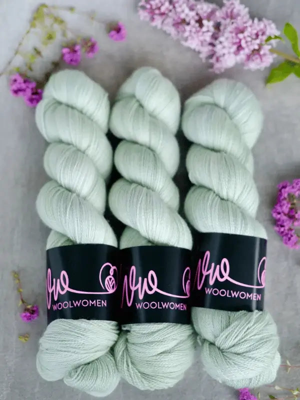Three skeins of pale mint green WoolWomen Merino Silk Lace yarn are arranged side by side on a light surface, surrounded by small clusters of purple flowers.