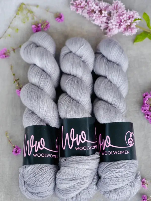 Three skeins of WoolWomen Merino Silk Lace yarn in soft light gray are arranged side by side on a gray surface, surrounded by delicate purple and pink flowers.
