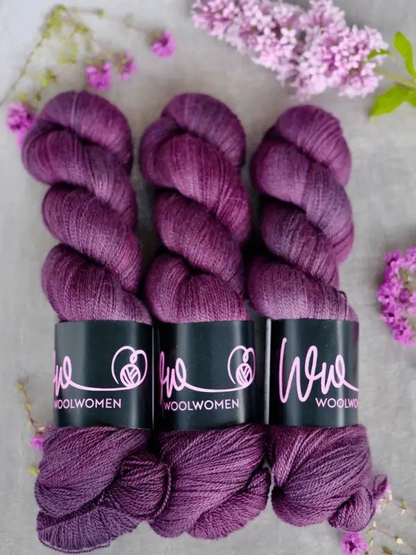 Three skeins of WoolWomen Merino Silk Lace in purple are lined up on a light surface, surrounded by lilac and purple flowers—perfect käsinvärjätty merinovilla pitsilanka for your next delicate project.