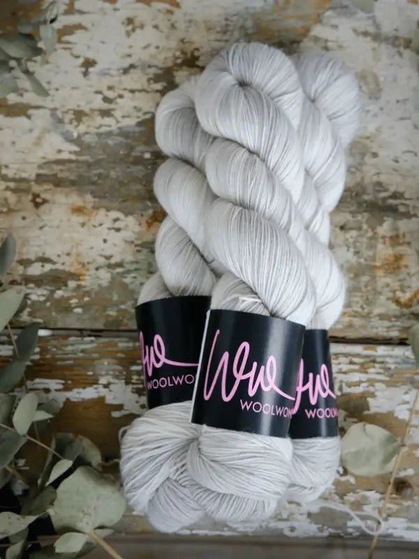 Two skeins of pale gray WoolWomen Merino Sock yarn rest on a rustic, weathered wooden surface, with dried leaves scattered at the edges.
