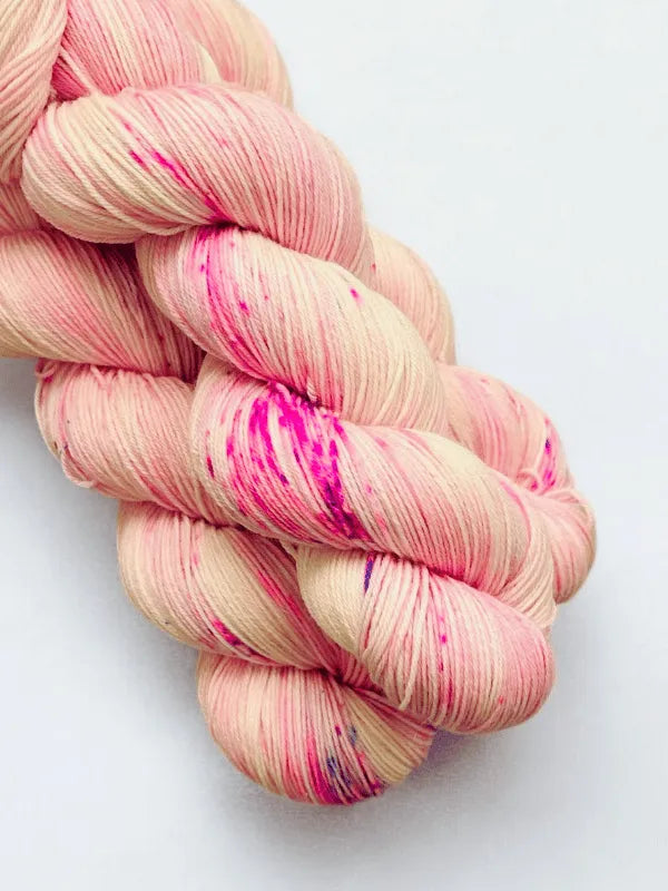 Two skeins of WoolWomen Merino Sock yarn twisted together, with a pale pink base, bright pink splashes, and subtle purple speckles, set against a white background.