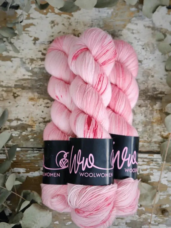 Three skeins of WoolWomen Merino Sock in pale pink and white with dark pink specks, each wrapped with a black label, are arranged on a rustic wooden surface surrounded by dried leaves.