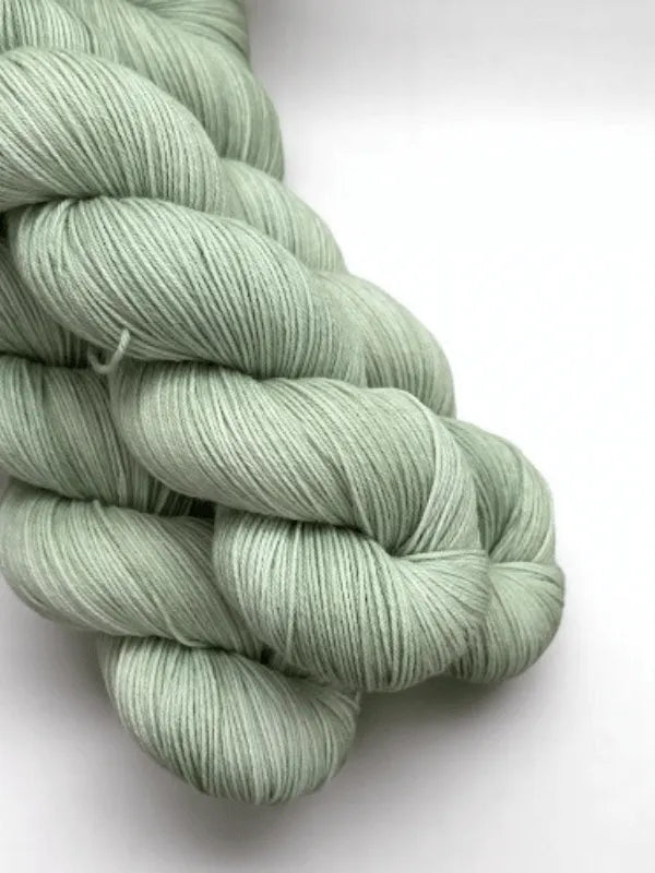 Three skeins of WoolWomen Merino Sock yarn in soft, pale green are neatly twisted against a white background. This smooth, fine superwash merinovilla yarn is ideal for delicate knitting or crochet projects.