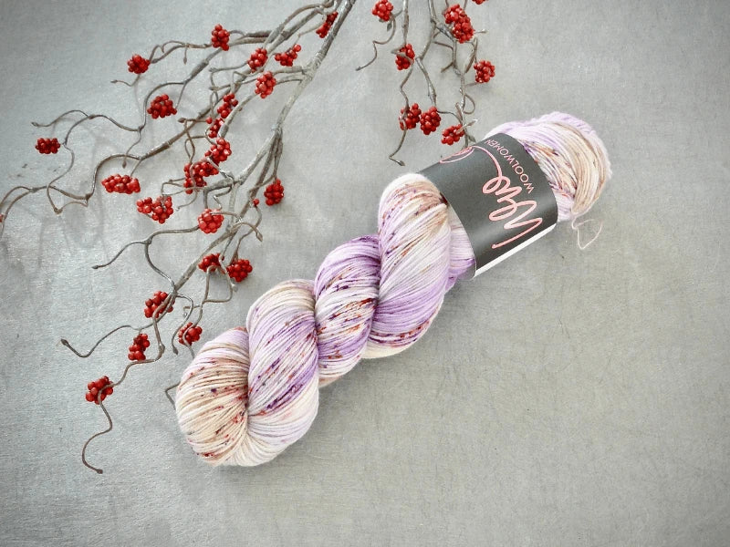 A skein of WoolWomen Merino Sock – Yhden Padan Ihme in light purple and cream speckles rests on a gray surface beside a branch with small red berries.