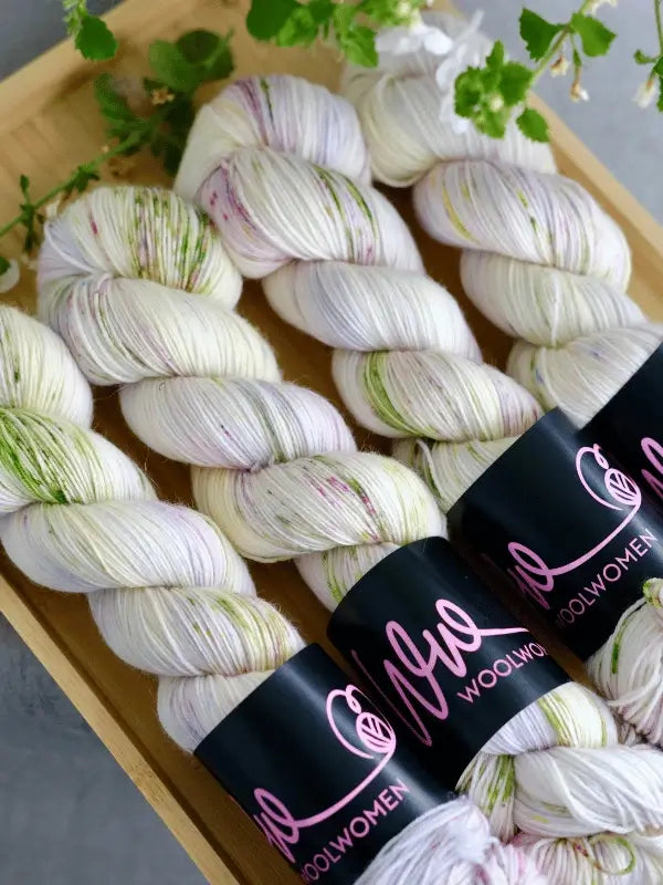 Three skeins of WoolWomen merino sock - Summer Collection yarn, white with green and pink speckles, are neatly arranged on a wooden tray. Each has a black label with pink text. Green plant leaves hang above the display.