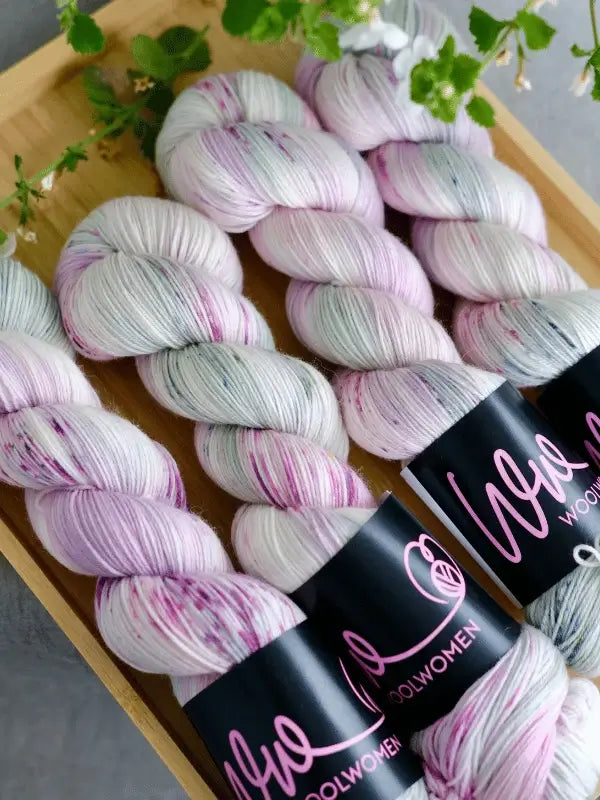 Four skeins of WoolWomen merino sock - Summer Collection yarn in hand-dyed white, pink, and gray with speckles are displayed on a wooden tray. Each skein features a black Wool Women label and green leaves accent the arrangement.
