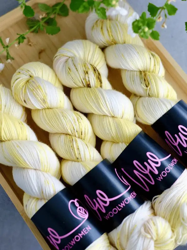 Four skeins of WoolWomen merino sock yarn from the Summer Collection, in pale yellow and white with subtle speckles, are displayed side by side in a wooden tray. Each has a black label with pink script; green leaves adorn one corner.
