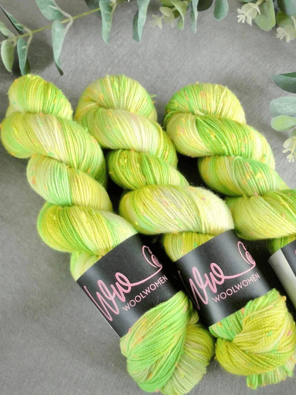 Three skeins of WoolWomen neon tweed yellow and green variegated superwash merinovilla yarn are arranged on a gray surface, framed by green leaves at the top and right edges.