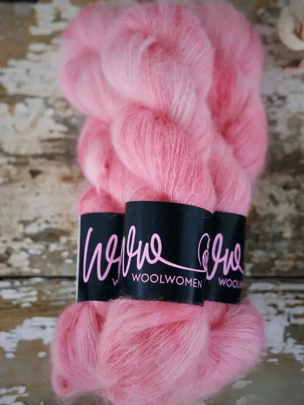 Two skeins of fluffy, pink yarn with black labels reading "WoolWomen Silk Mohair" are placed on a rustic, weathered wooden surface with white paint chipping off. This ylellinen neulelanka is a käsinvärjätty silkki-mohair-sekoitus, adding both luxury and softness to your next knitting project.