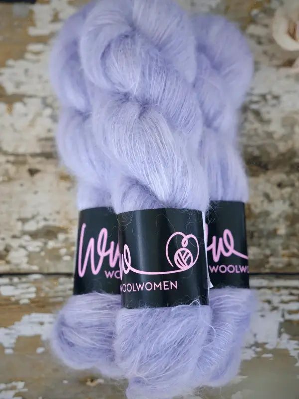 Two skeins of pale lavender WoolWomen Silk Mohair yarn with black labels featuring WOOLWOMEN in pink script, resting on a distressed wooden surface. This luxurious silk mohair yarn is käsinvärjätty mohair, ideal for crafting elegant and ylellinen neulelanka projects.