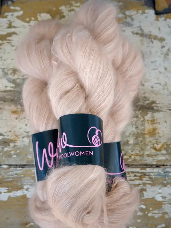 Two skeins of soft, pale pink WoolWomen Silk Mohair yarn with distinctive black and pink labels rest on a distressed wooden surface. This käsinvärjätty silkkimohairlanka is perfect for creating ylellinen neulelanka projects that exude elegance and warmth.