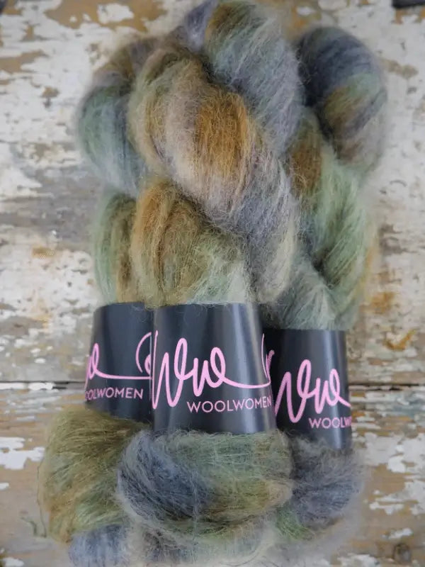 Three skeins of WoolWomen Silk Mohair—soft, multicolored yarn in käsinvärjätty shades of green, grey, and brown—are wrapped with black labels reading WOOLWOMEN and arranged on a rustic, weathered wooden surface.