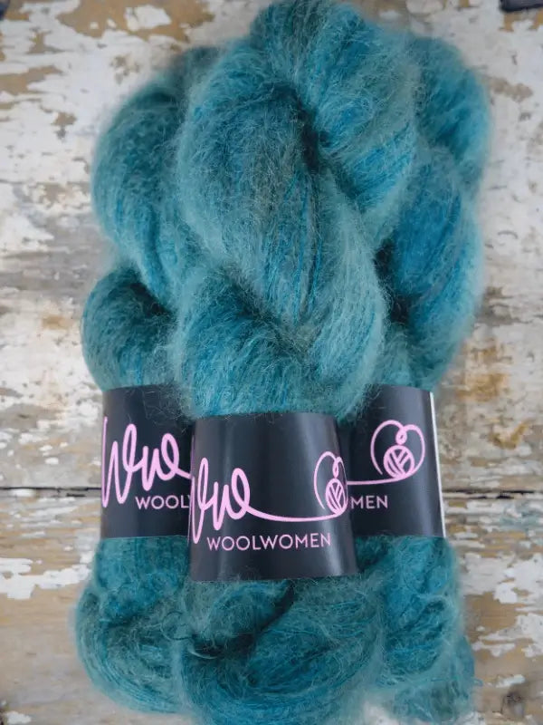 Two skeins of teal blue fluffy yarn labeled WoolWomen Silk Mohair, wrapped with black bands and pink writing, are arranged on a distressed wooden surface. This luxurious silk-mohair yarn is perfect for your next project, offering the softness and sheen that only an ylellinen neulelanka can provide.