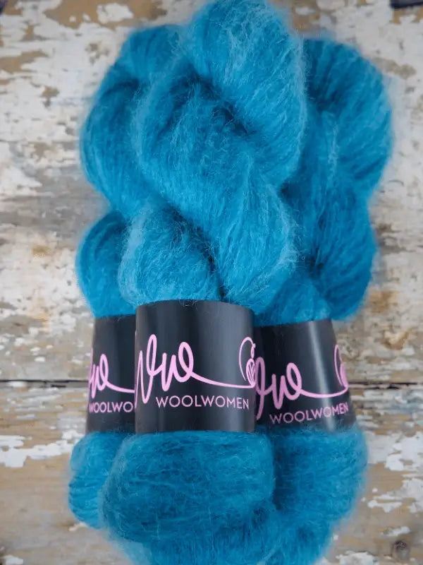 Two skeins of vibrant blue WoolWomen Silk Mohair yarn with black labels rest on a weathered, rustic wooden surface. This ylellinen neulelanka brings luxurious texture and brilliant color to your next knitting project.