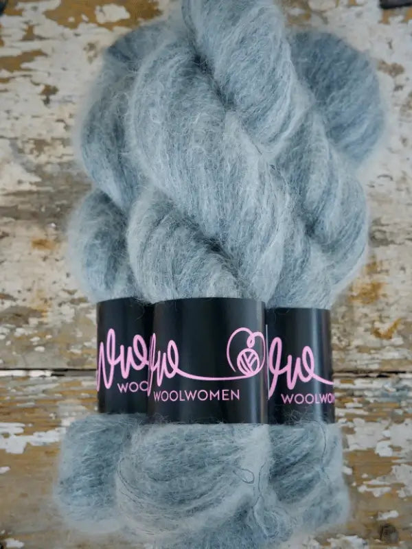 Two skeins of WoolWomen Silk Mohair, in a soft light blue shade, are twisted and wrapped with black labels featuring the brand name in pink. The luxurious silk-mohair yarn rests on a rustic, weathered wooden surface, highlighting its delicate sheen and sumptuous texture.