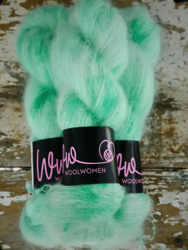 Three skeins of mint green WoolWomen Silk Mohair yarn, featuring a fuzzy texture and wrapped in black labels with "WoolWomen" in pink, rest on a rustic wooden surface. This käsinvärjätty mohair brings a touch of luxury to your projects—perfect for knitters seeking a truly ylellinen neulelanka.