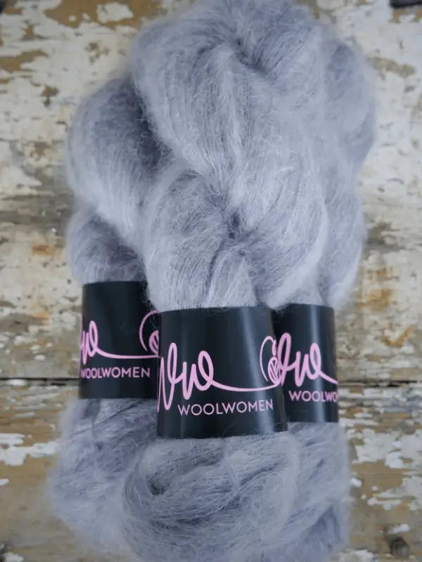 Three skeins of light gray, fluffy yarn labeled WoolWomen Silk Mohair with black and pink bands are arranged on a rustic, weathered wooden surface. This luxurious silk mohair yarn is perfect for your next project, adding exceptional softness and elegance to any creation.
