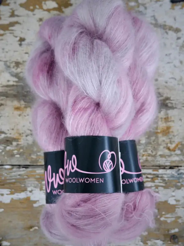 Three skeins of fluffy, light pink and white WoolWomen Silk Mohair yarn with black labels reading WOOLWOMEN are stacked on a rustic, distressed wooden surface.