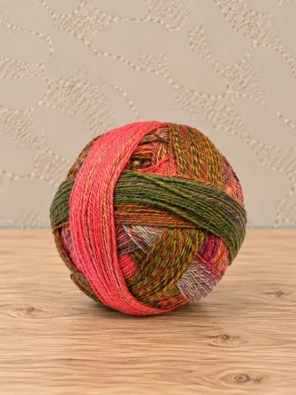 A round ball of Schoppel wolle Zauberball crazy yarn in variegated shades of green, brown, pink, and orange.