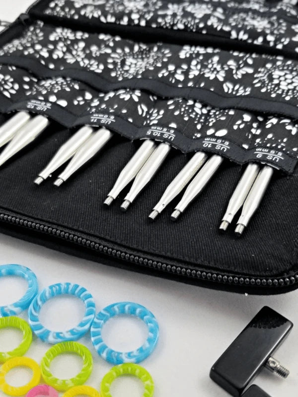 A black zippered case with the ChiaoGoo TWIST Complete - Set is shown on a white surface, featuring patterned slots, colorful stitch markers, and a black tightening tool.