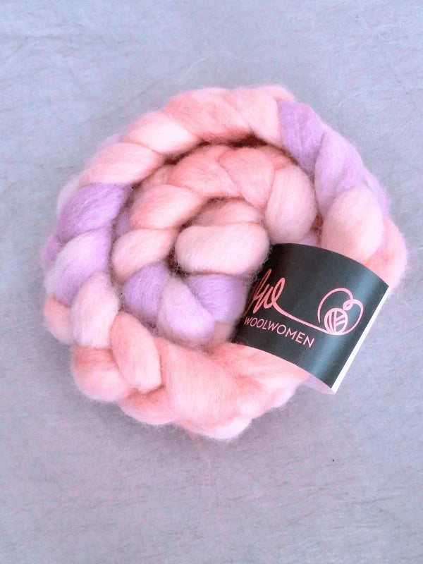A coiled braid of soft, thick WoolWomen Suomalainen Kehruuvilla käsinvärjätty wool roving in pastel pink and lavender, with a black and pink Wool Women label, displayed on a light gray surface.