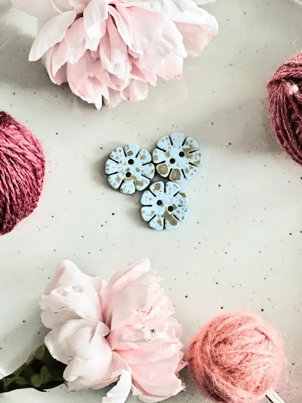 Two pink flowers and two balls of pink yarn frame four blue Värikkäät kukkanapit 20mm (17700), arranged in a circle on a light, speckled surface for a rauhallinen ja yksinkertainen composition.