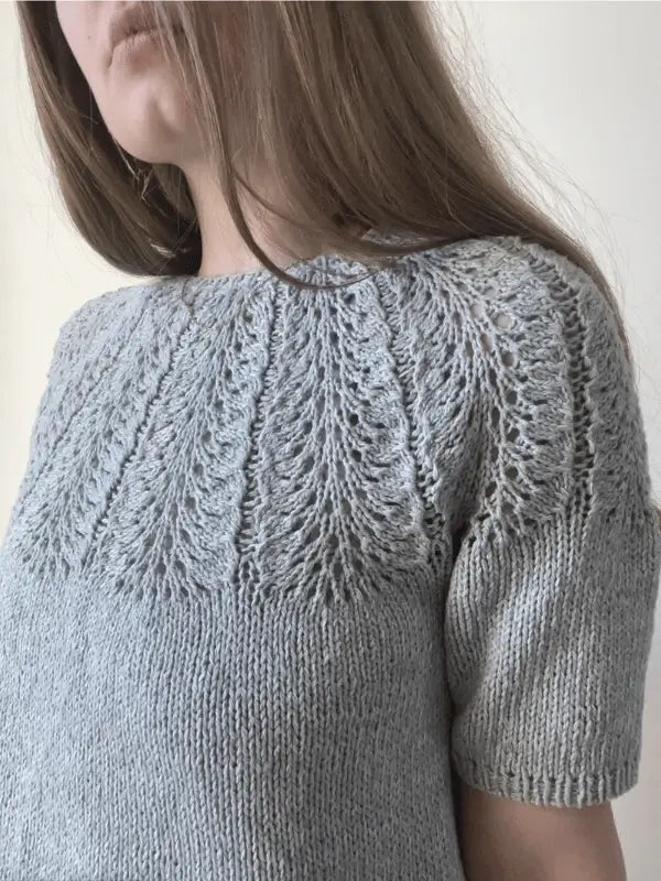 A woman with long brown hair wears a light gray Le Knit Palme Tee neuleohje, ENG, featuring a lace-like yoke and short sleeves. The background is plain and light-colored.