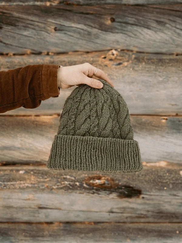 A hand in a brown sleeve holds the green, cable-knit Ronja Hakalehto Kiertoteitä beanie against a rustic wooden wall—perfect inspiration for neuleohjeet from Pohjois-Suomi.