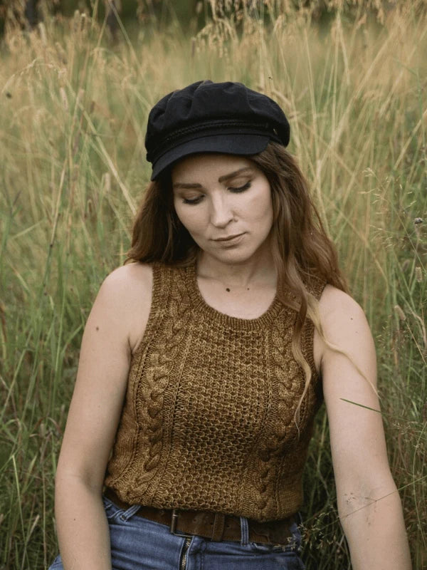 A woman with long brown hair, resembling Ronja Hakalehto, sits calmly in tall grass wearing a sleeveless knit top and black newsboy cap—an image inspired by "Ronja Hakalehto, Kiertoteitä.