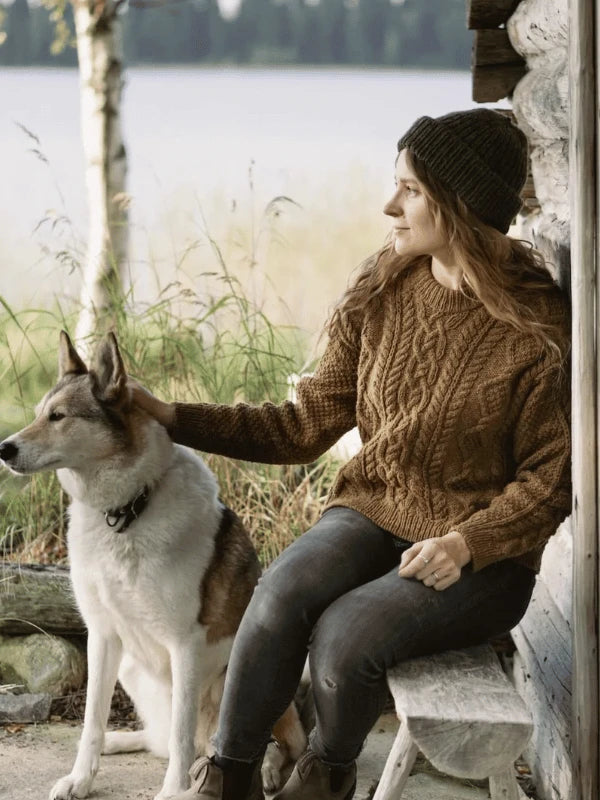 Wearing a brown sweater and knit hat inspired by Ronja Hakalehto, Kiertoteitä, a woman sits on a wooden bench outside a rustic cabin, petting her large dog as tall grass and a lake stretch out in the background.