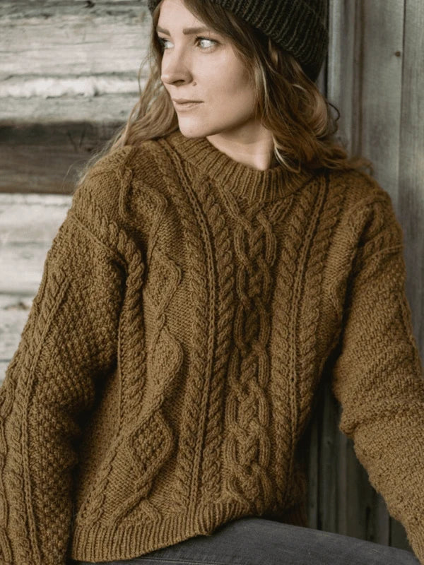 A woman in a brown cable-knit sweater and dark beanie, inspired by Ronja Hakalehto’s "Kiertoteitä," sits thoughtfully before a rustic wooden wall.