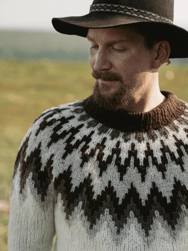 A bearded man in a black wide-brimmed hat and a black-and-white zigzag sweater inspired by Ronja Hakalehto's Kiertoteitä stands outdoors with the grassy landscape of Pohjois-Suomi in the background.
