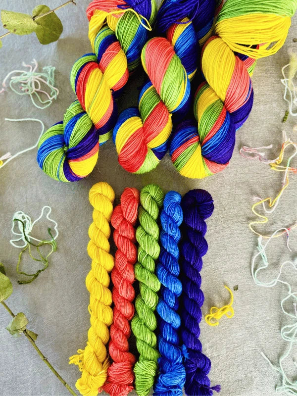 Six skeins of brightly colored yarn, including WoolWomen minisetit in tehostevärit, are arranged at the top, with six smaller merinovilla lanka skeins in rainbow colors below, all on a gray surface scattered with yarn.