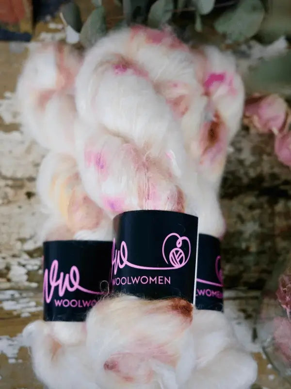 Three skeins of WoolWomen Fluff Mohair yarn—fluffy white with pink and brown accents and wrapped in black labels—are stacked on a rustic wooden surface.
