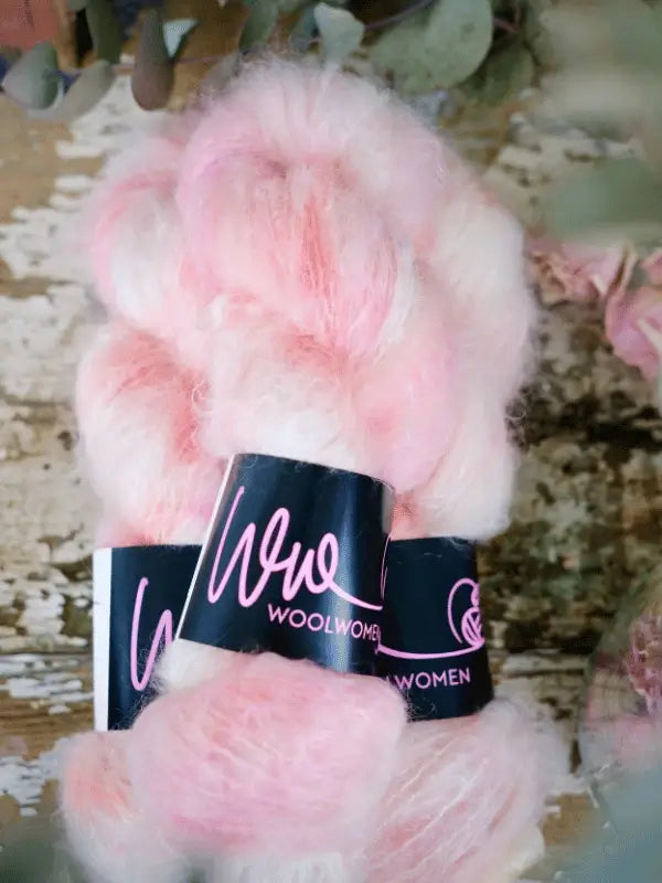 Two skeins of WoolWomen Fluff Mohair yarn in soft, fluffy pink and white are displayed on a rustic wooden surface. Their labels feature WOOLWOMEN in pink and white lettering on a black background.