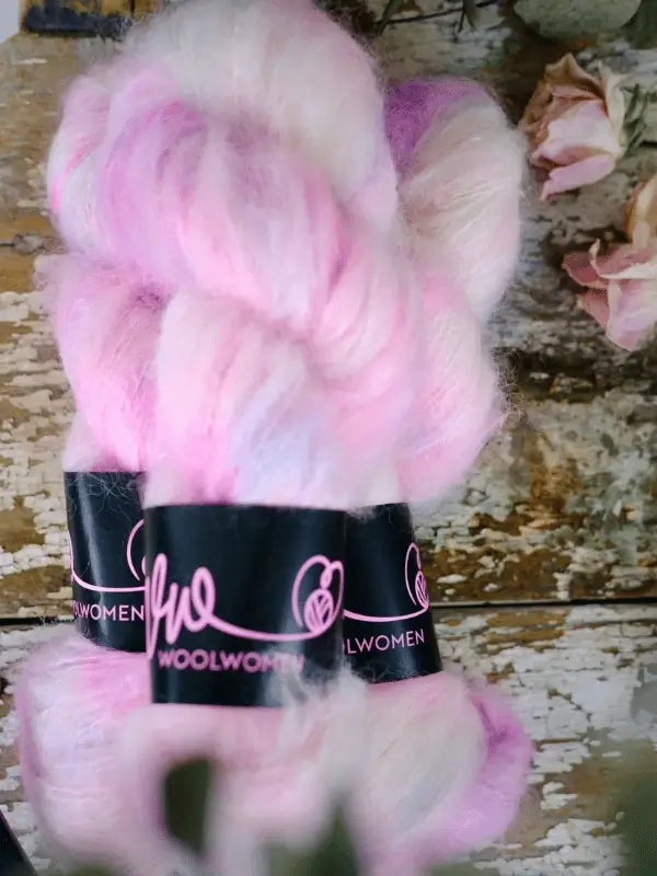 Three skeins of WoolWomen Fluff Mohair in pastel pink and white rest on a rustic wooden surface, with dried pale pink roses in the background.