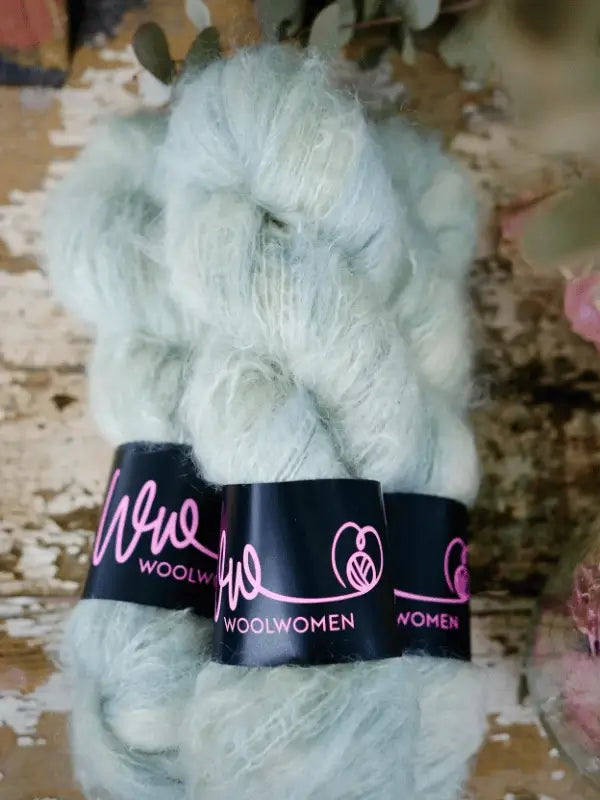 Two skeins of pale green WoolWomen Fluff Mohair yarn with black labels featuring pink text, displayed on a rustic wooden surface.