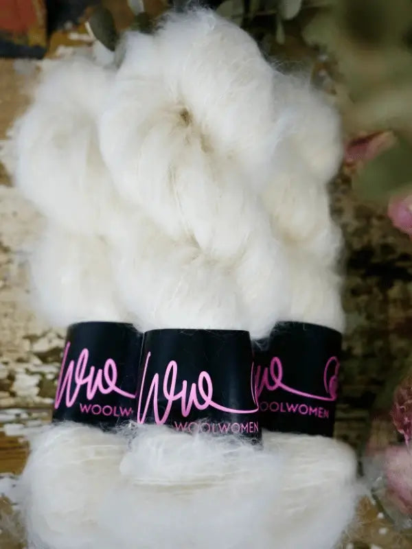 Three bundles of WoolWomen Fluff Mohair are stacked together, each wrapped with a black label featuring pink cursive text. The background is blurred with rustic and floral elements.
