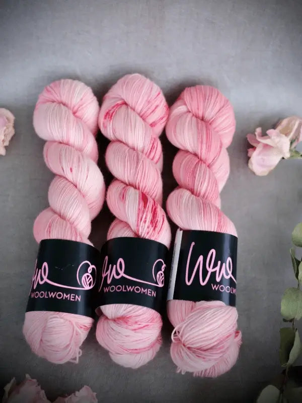Three skeins of WoolWomen Merino Sock yarn in pale pink with darker pink speckles, wrapped in black WoolWomen labels, are arranged side by side on a gray surface accented with flowers.