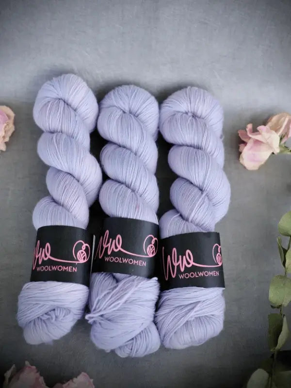 Three skeins of soft, pastel lavender WoolWomen Merino Sock yarn rest side by side on a gray surface, with pale pink flowers in the image corners.