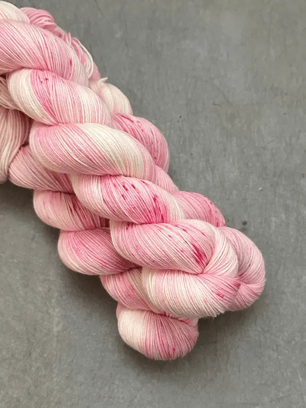 Two skeins of WoolWomen Merino Sock yarn in pale pink and white with subtle red speckles, twisted together and displayed on a gray surface.