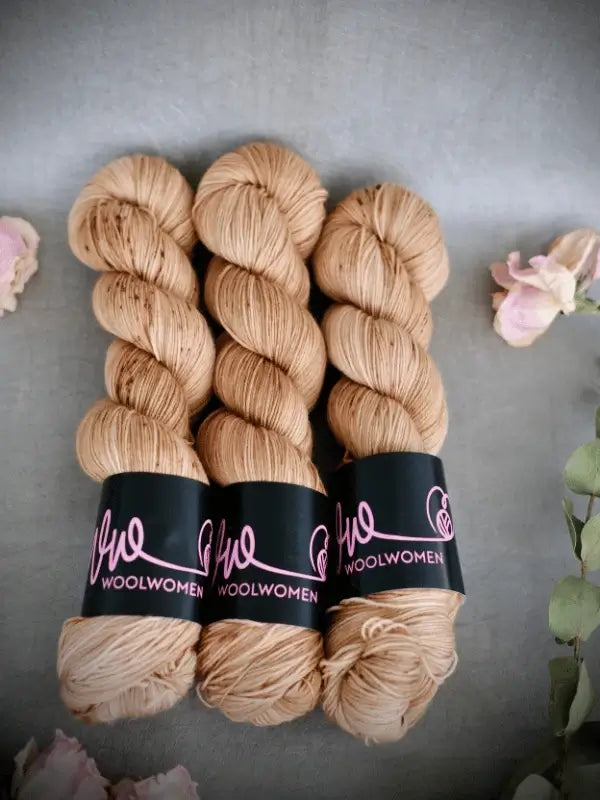 Three skeins of WoolWomen Merino Sock yarn in light brown, each with a black WOOLWOMEN label, are arranged side by side on a gray surface, surrounded by softly blurred pink and green flowers.